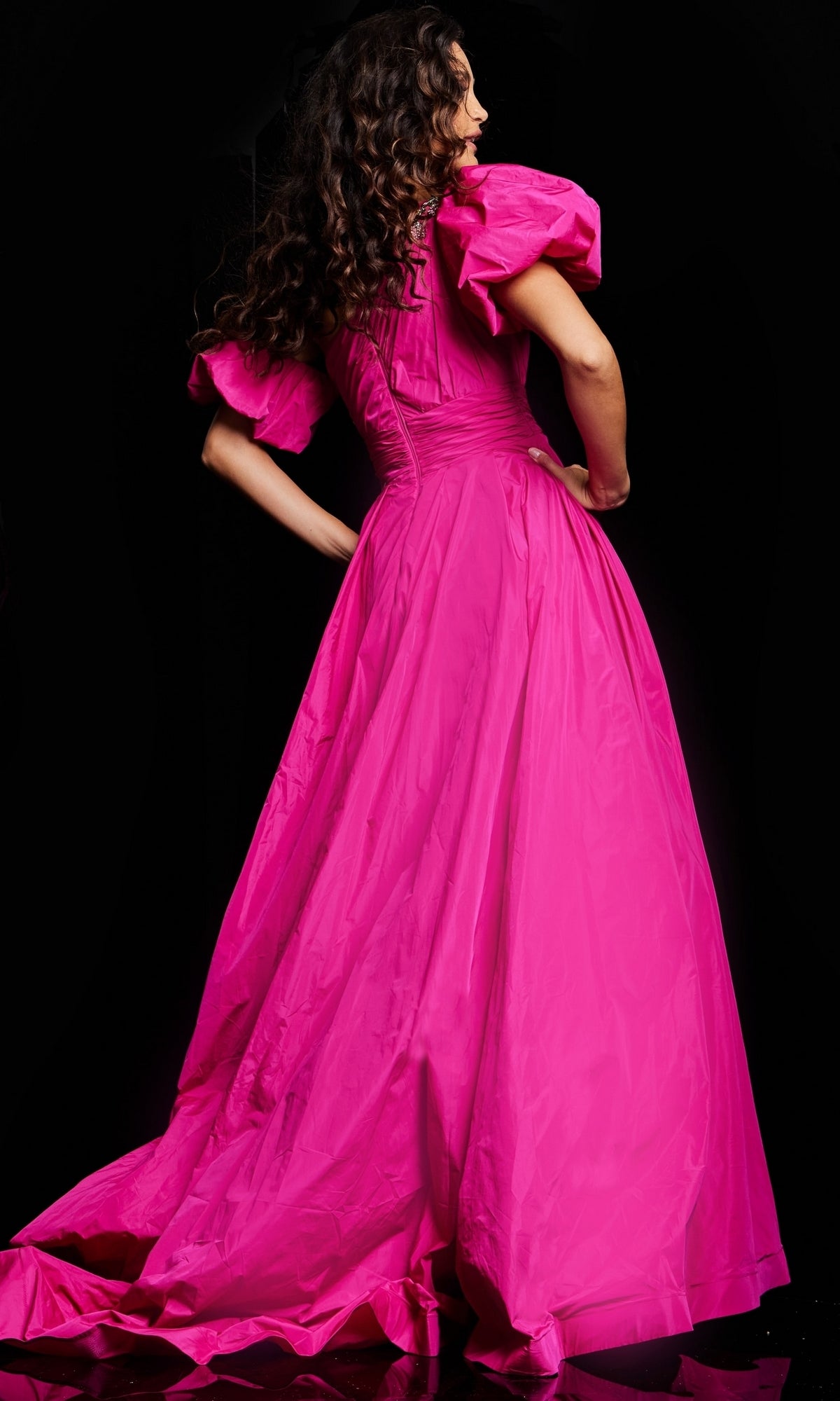 A person with long, wavy hair faces away, wearing the Long Prom Dress 36872 by Jovani—a bright pink ball gown with puffed short sleeves—set against a black background.